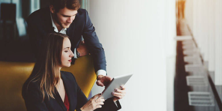 Lady sitting in a mustard-coloured armchair looking at her offshore investment portfolio while her financial adviser points to something on the i-Pad that she’s holding.